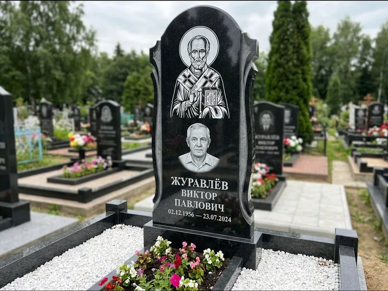 Памятник с гравировкой иконы: стела с образом в полный рост на обороте — гравировка образа Матроны Московской, низкие кусты спиреи в цвету, работа «Лебедева» в Альметьевске